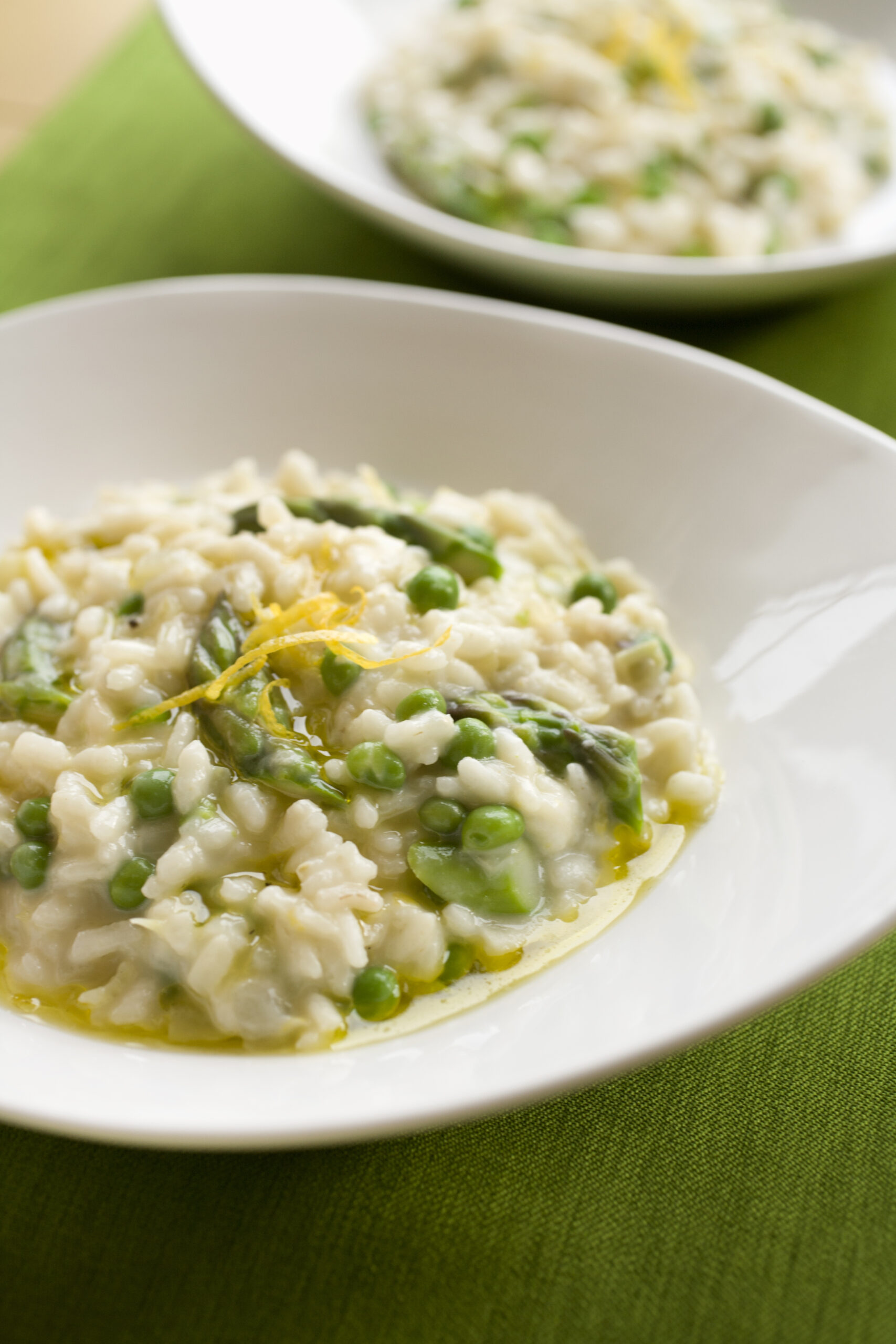 "Two servings of risotto with fresh green peas, asparagus spears and garnished with grated lemon zest.  Served in modern, white bowls on a bright green table runner."